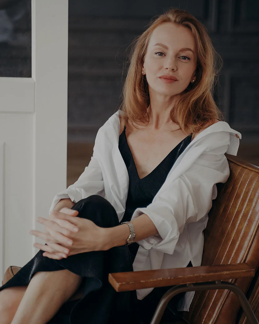 A middle-aged woman with light reddish-brown hair sits in a wooden chair, wearing a white button-up shirt over a black top and dark pants or a skirt. Her hands rest on her lap, and she wears a silver watch on her right wrist. The background features a white door and a dark gray wall. Soft, even lighting highlights the details. - Eyelid Surgery in Dunedin, FL